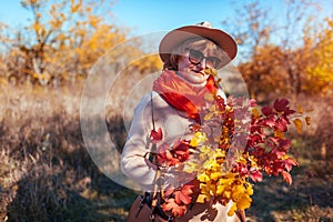 Middle-aged woman holding bouquet of autumn branches with yellow and red leaves. Lady walking in forest