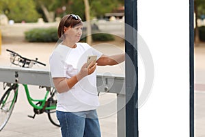 Middle-aged woman holding smartphone is paying the urban bicycle rental service in the European city at rental station