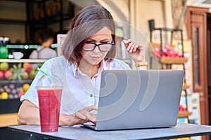 Middle-aged woman with a laptop at a table in an outdoor cafe with fresh juice