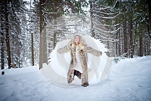 A middle-aged woman in a large warm fur coat and white angel wings in a winter forest with snow and snowdrifts. Fairy angel in a