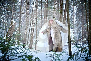 A middle-aged woman in a large warm fur coat and white angel wings in a winter forest with snow and snowdrifts. Fairy angel in a