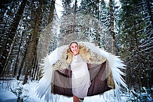 A middle-aged woman in a large warm fur coat and white angel wings in a winter forest with snow and snowdrifts. Fairy angel in a