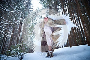 A middle-aged woman in a large warm fur coat and white angel wings in a winter forest with snow and snowdrifts. Fairy angel in a