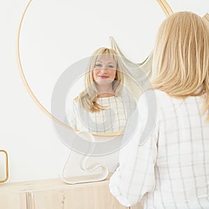 Middle-aged woman looks at herself. Mature beautiful blonde woman with long hair admires reflection