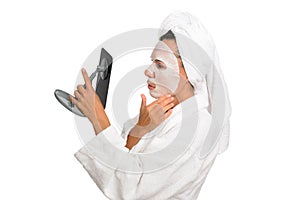 A middle-aged woman looks in the mirror and straightens a leafy hygenic mask on her face, wearing a white bathrobe. photo