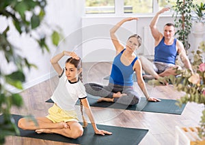 Middle-aged woman, man, preteen girl and boy warming up and doing side bends while sitting cross-legged before family