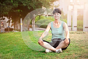 Middle aged woman meditating on the grass in public park outdoors in summer sunny morning