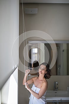 Middle-aged woman opening white roller blinds on window