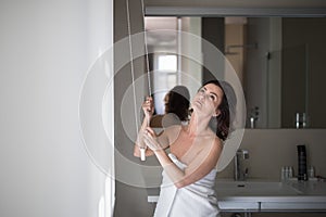 Middle-aged woman opening white roller blinds on window