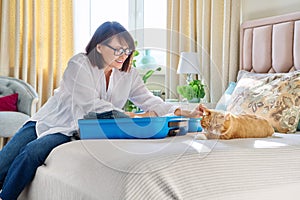 Middle-aged woman packing suitcase, sad cat lying on the bed