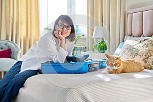 Middle-aged woman packing suitcase, sad cat lying on the bed