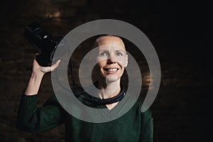 A middle-aged woman photographer holding a camera, smiling, and facing the camera against a dark background