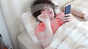 Middle-aged woman in pink pajamas lying in a bed watching phone