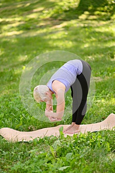 A middle-aged woman practices yoga outdoors, in the Standing Forward Bend pose. Uttanasana.