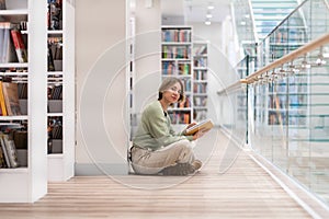 Middle-aged woman reader sitting on floor with book in hands in modern public library