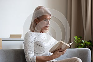 Middle aged woman reading book relaxing on sofa at home