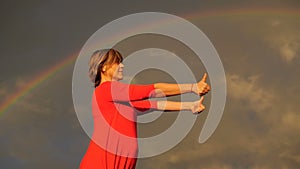 Middle aged woman in red dress shows thumbs and smile during rainbow.