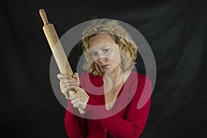 Middle-aged woman with red sweater threatens with a wooden rolling pin on dark background