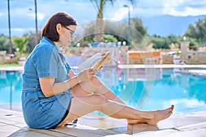 Middle aged woman resting near outdoor pool reading book