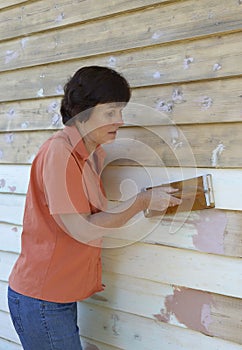 Middle-aged Woman Sanding Weatherboards on House.