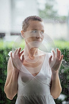 A middle-aged woman with short hair looks out from behind wet glass. Reflections, rethinking life concept.