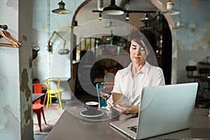 A middle-aged woman sits in a cafe, drinks coffee and works at a computer. Woman holding a phone