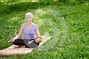 A middle-aged woman sits in a lotus position in a park,holds a singing bowl in her hands during a meditation session.