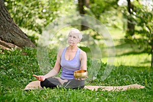 A middle-aged woman sits in a lotus position in a park,holds a singing bowl in her hands during a meditation session.