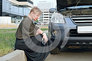 Middle-aged woman sits on the side of road in front of broken-down car with an open hood