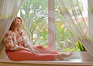 A middle-aged woman sits on the windowsill and looks thoughtfully out the window