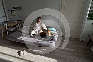 Middle-aged woman sitting on bed with dog in morning at home, enjoying living alone with beloved pet