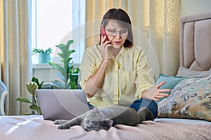 Middle-aged woman sitting on bed with laptop, cat talking on phone