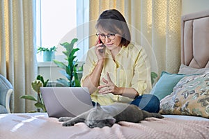Middle-aged woman sitting on bed with laptop, cat talking on phone