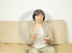 Middle aged woman sitting on the couch at home, holding glas of fresh water and taking medication.