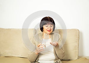 Middle aged woman sitting on the couch at home, holding glas of fresh water and taking medicine.