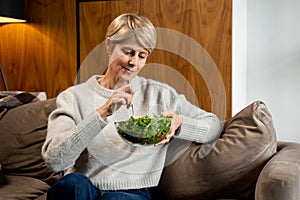 Middle-aged woman is sitting and resting at home on the couch, eating a green salad. A woman eating healthy food.