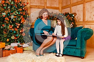 A middle aged woman and a six year old girl with dark hair are sitting on a sofa reading a book for Christmas