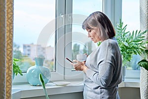 Middle-aged woman with smartphone, cup of coffee, at home near window