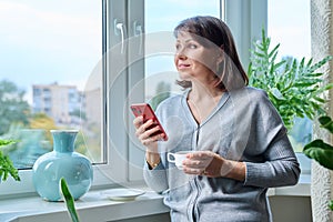 Middle-aged woman with smartphone, cup of coffee, at home near window