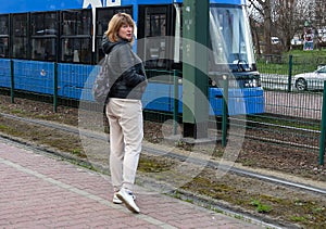 Middle-aged woman of a sports type in a jacket and sweatpants stands at a tram stop next to a tram, urban spring landscape