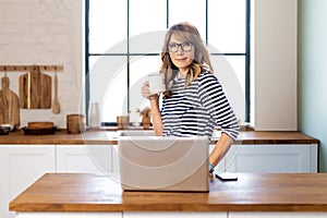 A middle-aged woman standing at the kitchen counter in the morning and using a laptop