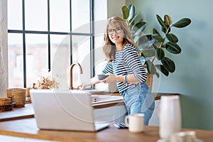 A middle-aged woman standing at the kitchen counter in the morning and using a laptop