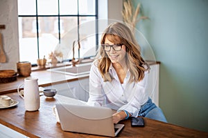 A middle-aged woman standing at the kitchen counter in the morning and using a laptop