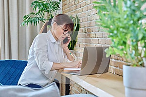 Middle-aged woman talking on mobile phone, sitting at desk at home with laptop