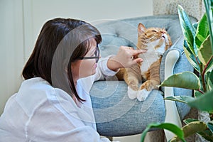 Middle aged woman touching ginger pet cat, home interior background