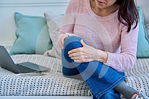 Middle-aged woman touching her sore knee joint with her hands, sitting at home