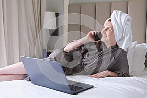 Middle-aged woman with towel on her head lies on bed, smiling and talking on the phone