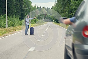 Middle-aged woman with a travel bag hitchhiking on the rural road in summer