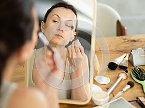 Middle-aged woman using brush during eye makeup sitting in front of the mirror