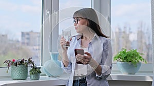 Middle aged woman using smartphone, drinking water standing near window